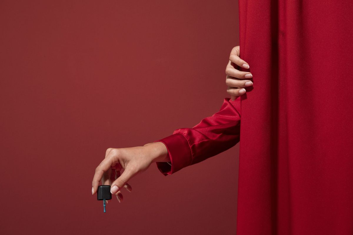 Manicured hand holding a nail polish bottle against a red curtain